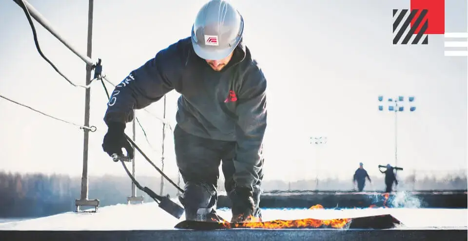 Un couvreur professionnel à genoux sur un toit, portant un équipement de sécurité et utilisant une torche pour installer une membrane de toiture, avec un ciel dégagé et des collègues au loin